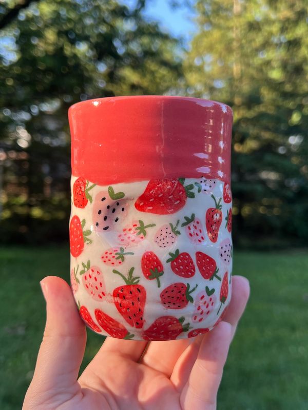 Handmade ceramic mug with hand-painted strawberry pattern and red glaze rim, held outdoors.