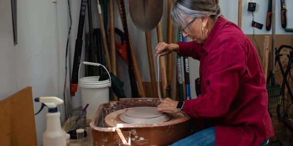 An elderly woman sculpts clay on a pottery wheel in a workshop.