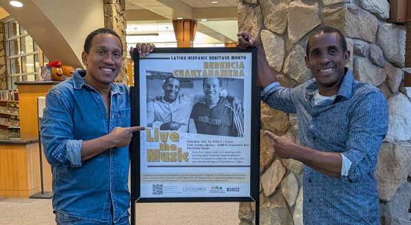 Two men smiling and pointing at a music event poster indoors.