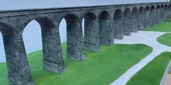 Model of a stone aqueduct on a grassy landscape with a winding path.