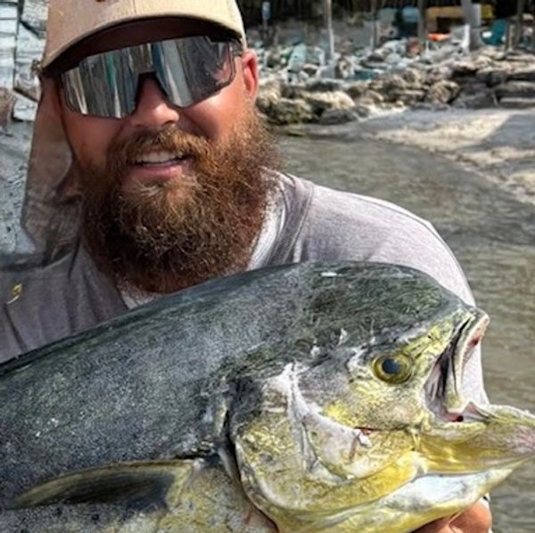 Bearded man proudly holding a large fish by the water.