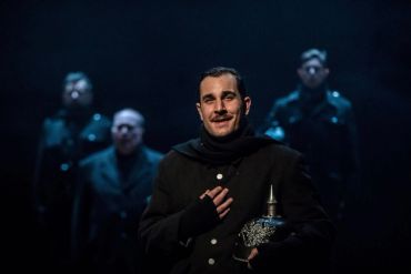 Man in dark costume on stage with three blurred figures.