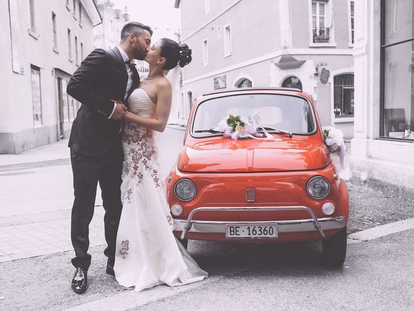 Bride and groom standing by a red car.