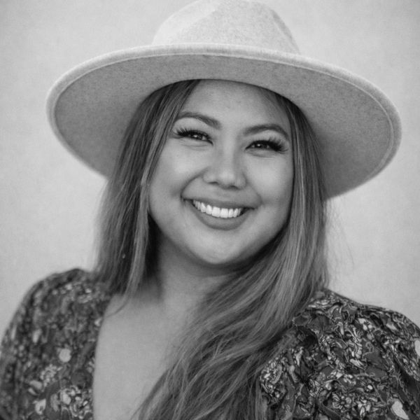 Smiling woman wearing a wide-brimmed hat and floral dress in a black-and-white portrait.