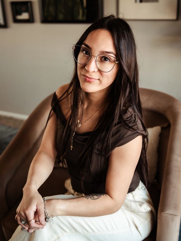 Young woman with glasses sitting on a chair, smiling gently.