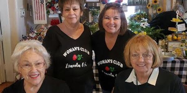Four women smiling, wearing Woman's Club Lincoln t-shirts indoors.