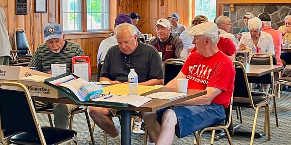 Seniors gathered indoors, engaging in reading and conversation around tables.