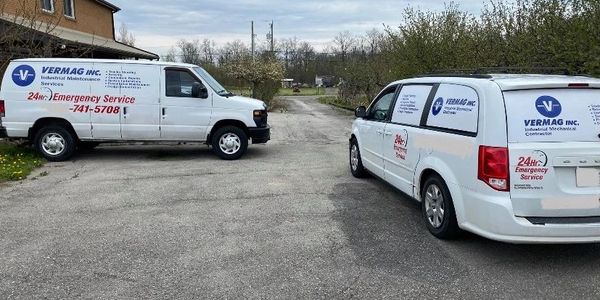 Two white company vans parked on a driveway near a brick building and trees.