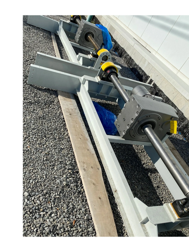 Industrial metal shaft assembly on gravel ground beside a building.