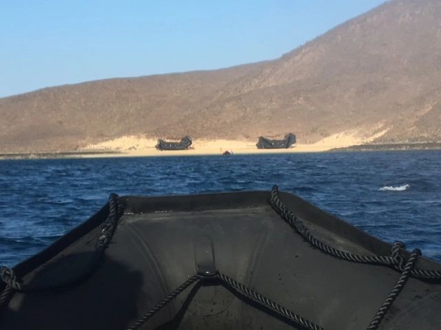 military zodiac headed toward beach with helicopters on the sand