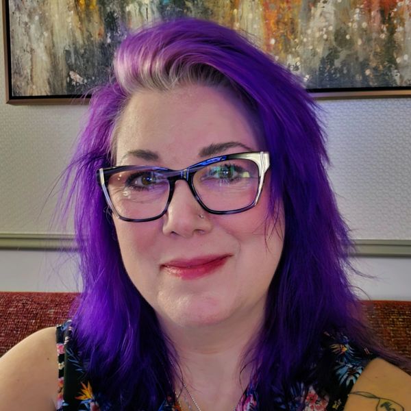 Woman with vibrant purple hair and glasses smiling indoors.