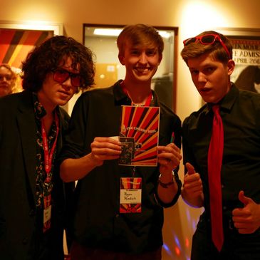 Three young men posing indoors, one holding a colorful booklet.