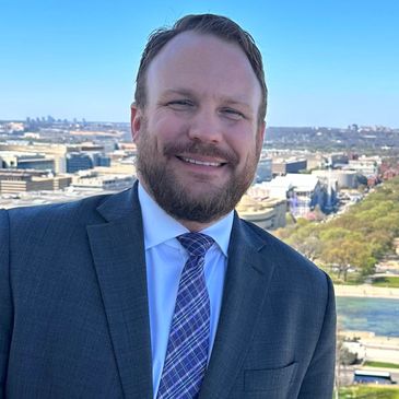 Smiling man in a suit with a cityscape background on a clear day.