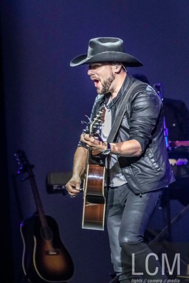 Man passionately playing guitar on stage wearing a cowboy hat and leather jacket.