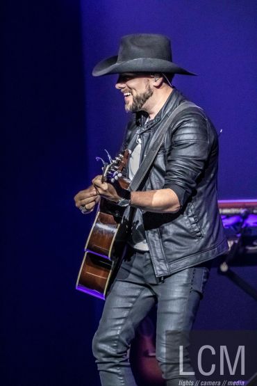 Musician in leather jacket and hat playing guitar on stage.