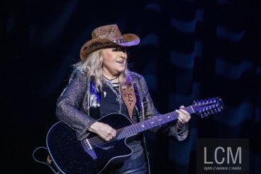 Musician playing a 12-string guitar on stage, wearing a cowboy hat.