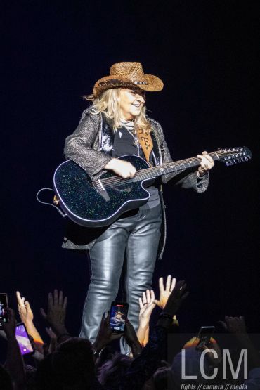 A musician in a cowboy hat plays guitar on stage with audience hands raised.