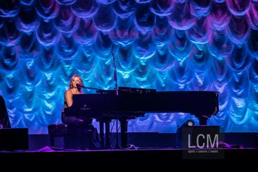 A woman playing piano on stage with blue and purple lighting.