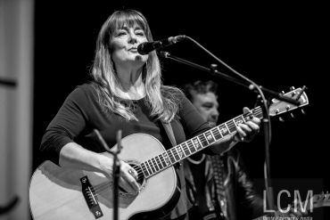 Female musician playing acoustic guitar and singing into a microphone on stage.