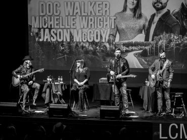Black and white image of a band performing on stage with a backdrop showing names.