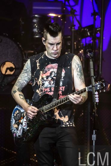 Musician intensely playing electric guitar on stage with tattoos and sleeveless rock shirt.