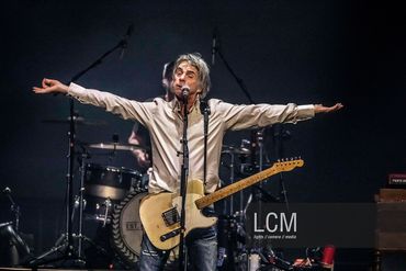 Singer playing guitar with arms outstretched on stage.