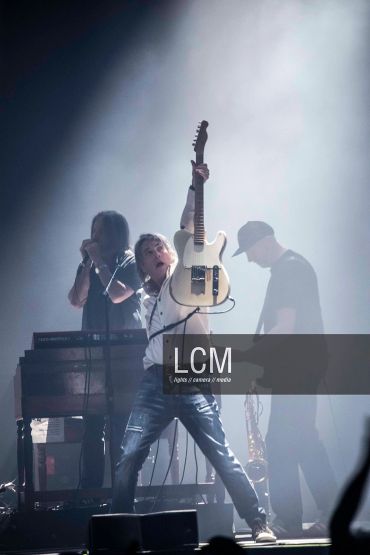 Musician raising guitar on stage with two band members behind.