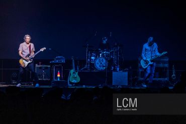 A three-member band performing live on stage with guitars and drums.