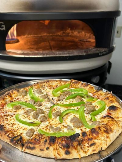Freshly baked pizza with green peppers and sausage in front of a pizza oven.