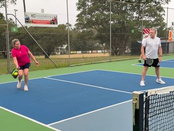 Two people playing pickleball on an outdoor court with a green and blue surface.