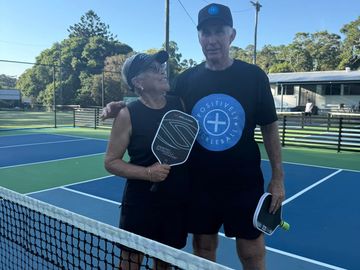 Two elderly people posing on a pickleball court with paddles, smiling and enjoying the game.