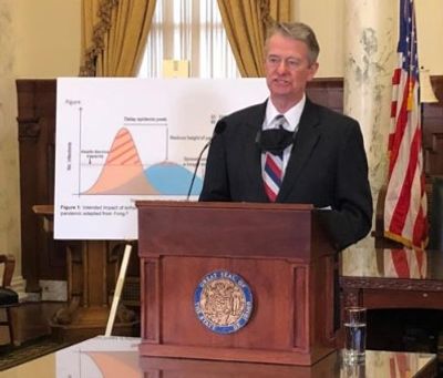 A man in a suit speaks at a podium with a chart behind him in a formal room.