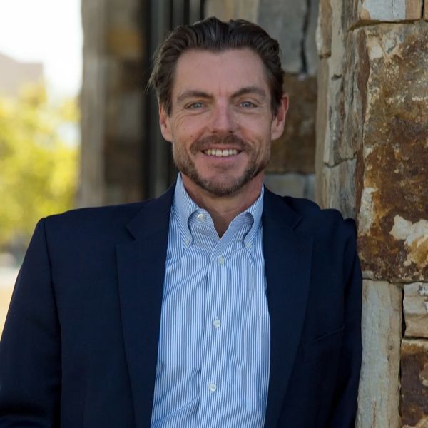 Man in a blue blazer smiles while leaning against a stone wall outdoors.