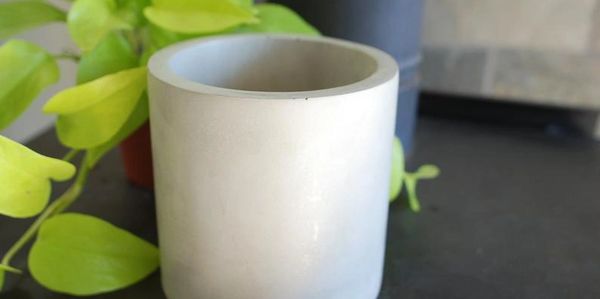 A simple white cylindrical ceramic pot with green plants in the background.