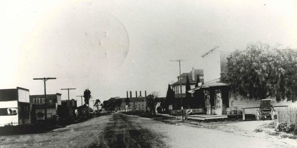 Historic photo of a dirt road in Los Alamitos surrounded by shops and houses.