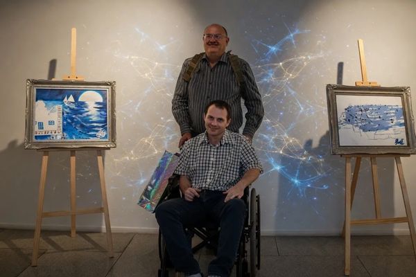 Two men, one in a wheelchair, pose in an art gallery with paintings on easels.