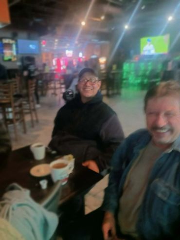 Two men smiling at a cafe table with drinks in a cozy setting.