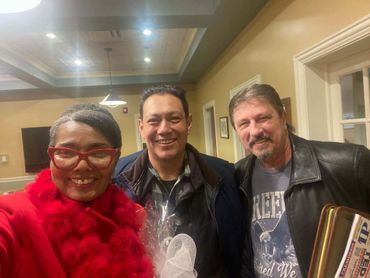 Three smiling people posing indoors, one wearing a red coat and glasses.