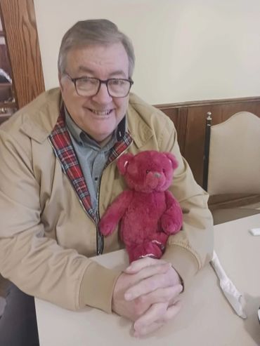 Man smiling with a red teddy bear on a table.