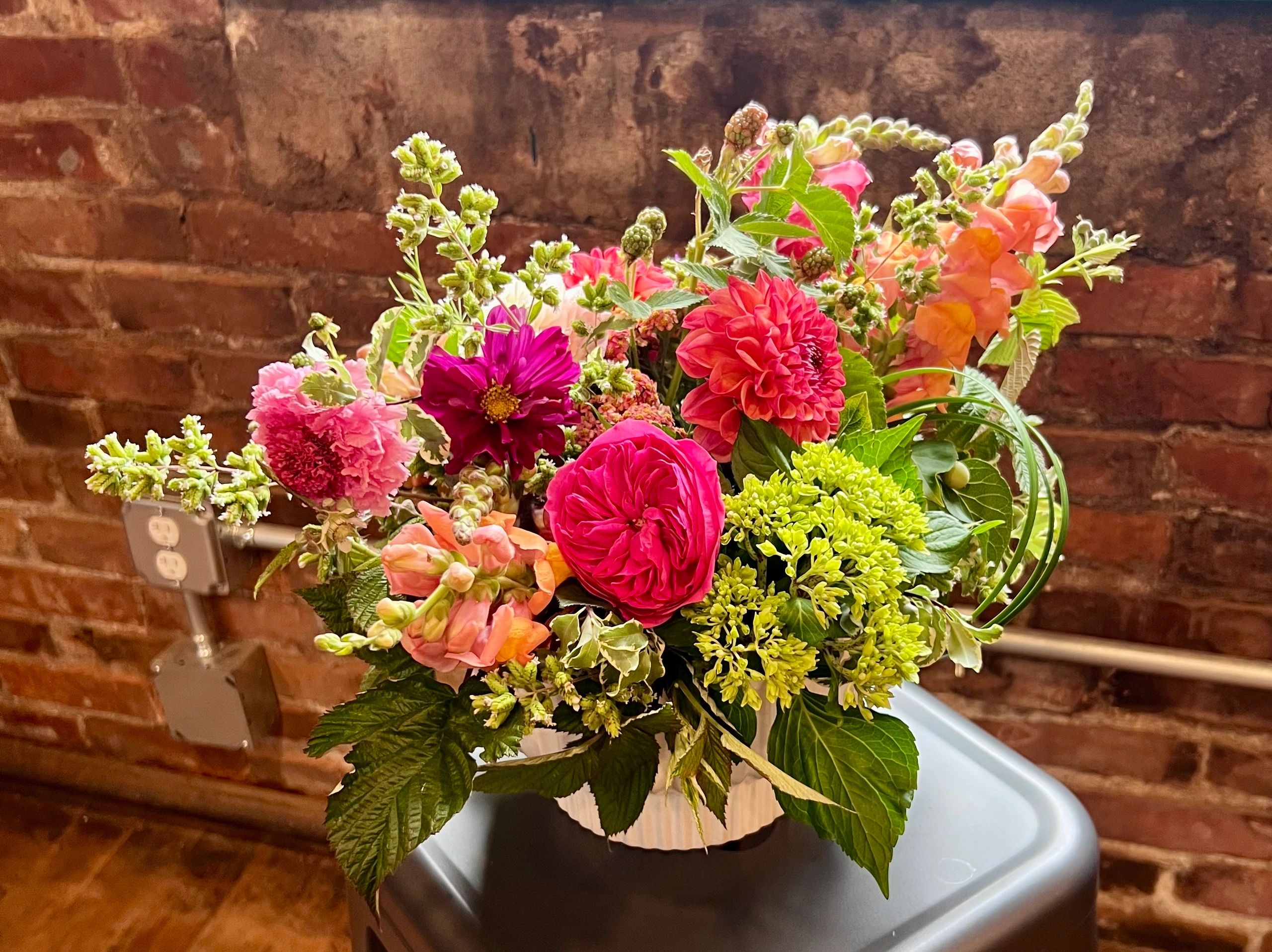 Vibrant mixed flower compote in a white vase against a rustic brick wall.