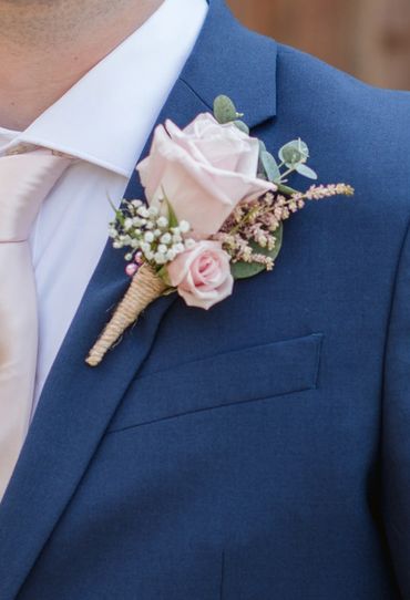 Smiling man in a blue suit with a pink boutonniere stands against a wooden background.
