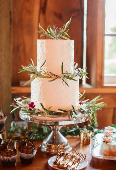 A rustic two-tier white cake adorned with greenery and purple flowers on a wooden table.