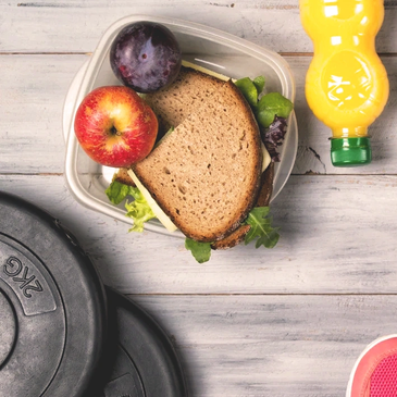 Healthy packed lunch with sandwich, apple, plum, and orange juice beside weights.
