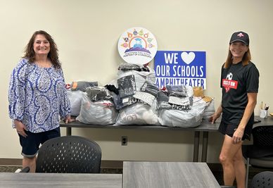 Two women stand beside a table with bags of donated clothing and a supportive school sign.