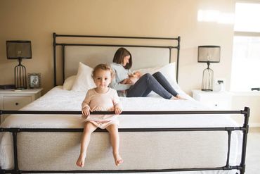 Toddler sitting on bed frame with mother in background.