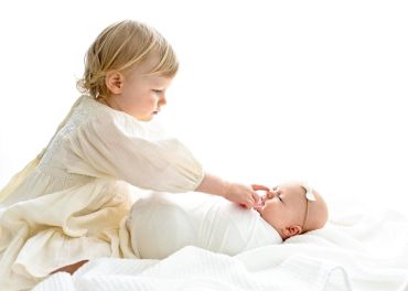 Toddler tenderly touches newborn baby swaddled in white.