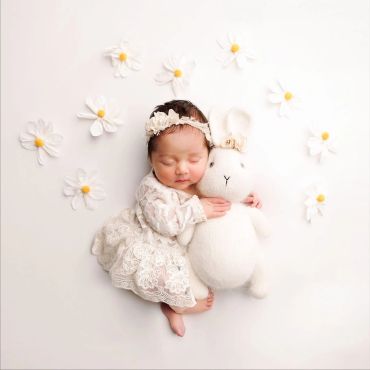 Sleeping baby in lace dress hugging a stuffed bunny surrounded by white flowers.