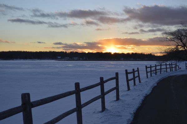 Snowy lake at sunset with wooden fence.