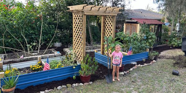 A young girl holding a shovel in a garden with raised beds and an arbor.