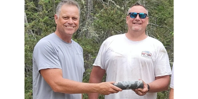 Two men outdoors smiling, holding a camouflaged object together.